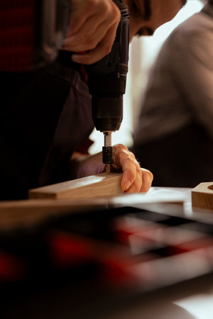 Manufacturer in carpentry shop using power drill to create holes for dowels in wooden board. Woodworking expert sinking screws into wooden surfaces with electric tool, doing precise drillingの写真素材