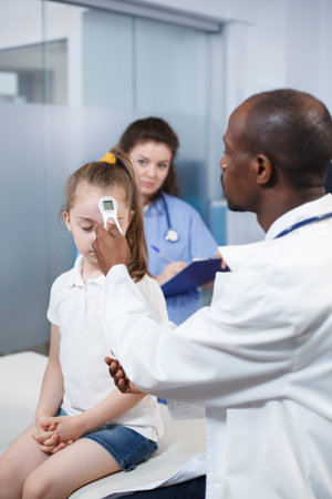 African American physician using thermometer on little girl to check fever in medical office. Doctor measures the temperature to find a diagnosis and gives treatment to the child in a cabinet.の写真素材