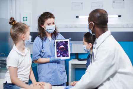Woman wearing blue scrubs holding a digital tablet presenting a virus cell picture in front of a girl and her mother, and the african american male physician. Expert treatment and education provided.の写真素材