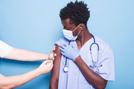 Caucasian person administering vaccine injection to African American nurse against blue background. Close-up of a doctor giving vaccination to black man, ensuring healthcare and covid 19 prevention.の写真素材