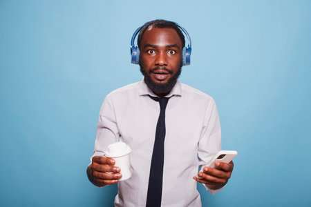 Shocked black man in white shirt looking at camera while holding cell phone for streaming music to wireless headphones. Young freelancer with a takeaway cup, having coffee and browsing internet.の写真素材