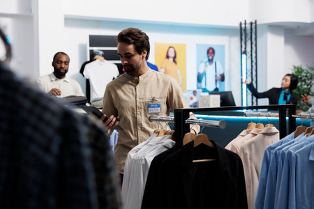 Clothing store manager using digital tablet and organizing merchandise to attract customers. Shopping center employee managing inventory and examining apparel hanging on rackの写真素材