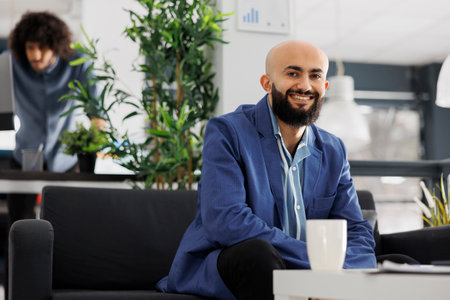 Smiling arab business entrepreneur looking at camera while sitting on couch in office. Start up company successful executive manager wearing suit portrait in coworking spaceの写真素材