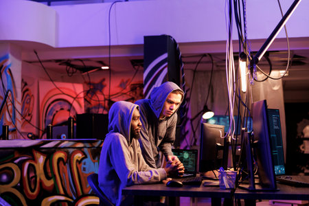 Hacker and crime partner working together and planning system breach in abandoned warehouse. African american and asian criminals checking ransomware on computer in hideoutの写真素材