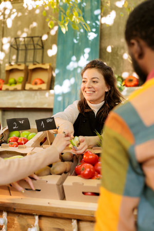 Smiling woman vendor offering customer to try out small piece of organic apple while selling fresh natural fruits and vegetables at harvest fair festival, selective focus. Tasting during shopping.の写真素材