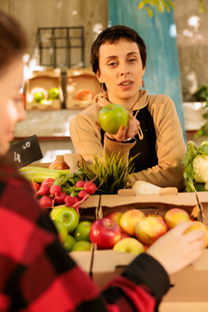 Local vendor wearing an apron and talking to customers while selling organic fresh apples at a farmers market stand. A young woman holds bio natural farming products grown locally.の写真素材