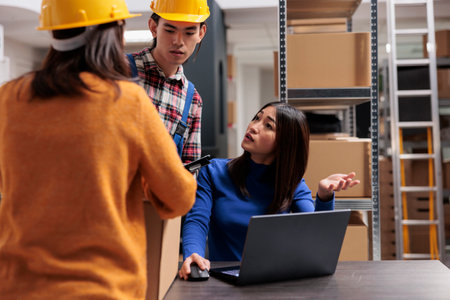 Asian warehouse workers team doing inventory tracking on laptop. Storehouse man and women employees managing delivery operations while working on computer in storage roomの写真素材