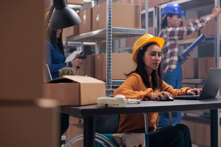 Asian postal service worker with physical disability using laptop in warehouse. Young woman wearing yellow protective hardhat in wheelchair tracking parcels shipment online on computerの写真素材
