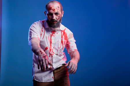 Portrait of evil reanimated zombie ready to attack and eat brains, infected by virus. Terrifying undead creature covered in blood and with rotten skin limping towards camera, studio backgroundの写真素材