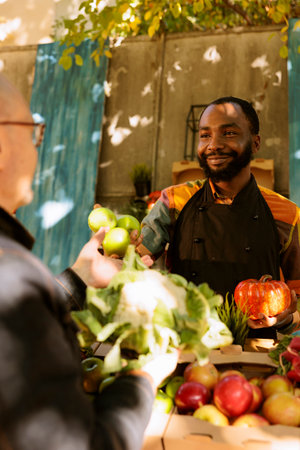 Cheerful farmer presenting locally grown apples on farmers market stand, small business organic products. African american man talking to senior customer, various fresh fruits and veggies.の写真素材