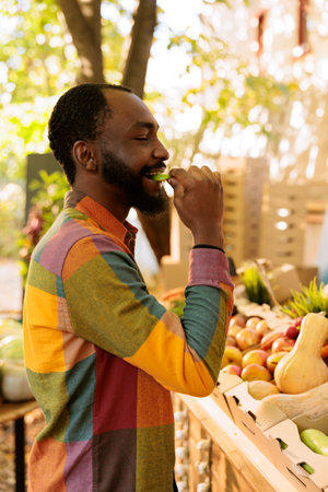 Portrait of smiling male individual tasting apple before buying bio produce, shopping for natural organic products at farmers market. Young black man enjoying food tasting fresh local produce.の写真素材