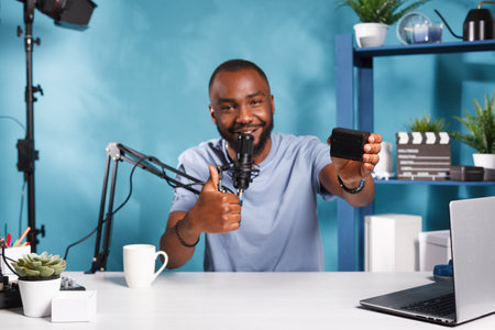 Smiling videographer recording camera gear positive review and showing thumb up gesture. Blogger live streaming and talking about photography equipment and holding accumulatorの写真素材