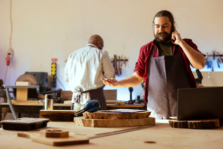 Smiling manufacturer discussing on telephone call with client inquiring about furniture assembling commission progress. Cheerful carpenter in assembly shop talking on mobile phone with customerの写真素材