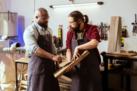 Carpenter holding timber block, brainstorming with coworker next steps in wood processing. Manufacturer in joinery and african american apprentice discussing what to do with piece of woodの写真素材