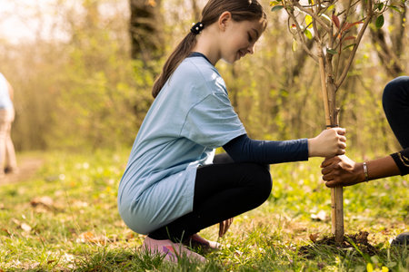 Little girl and her friend planting a tree in the ground, volunteering to the conservation project. Two teenagers taking action for reforestation, increasing vegetation in the forest, restore nature.の写真素材