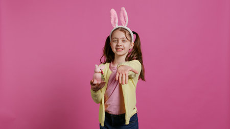 Lovely little girl with bunny ears sending air kisses in studio, presenting a stuffed pink rabbit toy for easter holiday. Smiling energetic preteen feeling positive and carefree. Camera B.の写真素材