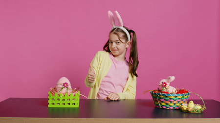 Happy schoolgirl with bunny ears showing thumbs up sign, feeling excited about easter holiday preparation. Cheerful child doing like okay sign and decorating baskets for festivity. Camera B.の写真素材