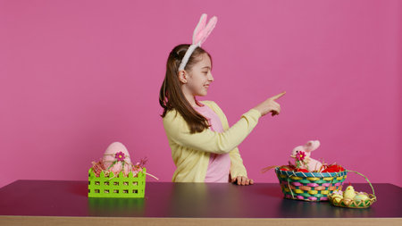 Adorable little kid indicating all directions in front of camera, pointing up, down, left and right while sitting at the table. Smiling lovely girl with bunny ears looking around. Camera B.の写真素材