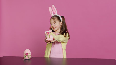 Smiling toddler with bunny ears showing her handmade easter ornaments, presenting a handcrafted decorated egg and rabbit. Young sweet schoolgirl holding festive decorations. Camera A.の写真素材