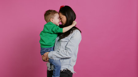 Adorable small boy hugging his mother and playing around, having fun against pink background. Young toddler posing with his mom holding him, laughing and being cheery. Camera B.の写真素材