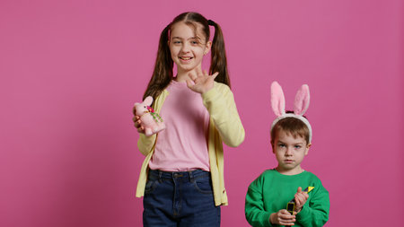 Cute brother and sister posing against pink background in studio, wearing bunny ears and playing with toys. Cheerful siblings feeling excited about easter, traditional spring holiday. Camera B.の写真素材