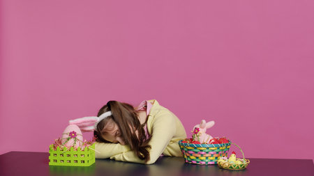 Exhausted little child decorating festive ornaments for easter sunday celebration, creating colorful cute arrangements with painted eggs. Tired girl yawning and falling asleep on the table. Camera A.の写真素材