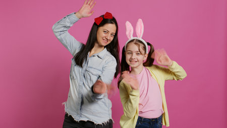 Smiling cheerful mother and little girl waving in front of camera, having fun and enjoying easter holiday celebration. Joyful schoolgirl posing with her mom in studio, saying hello. Camera B.の写真素材