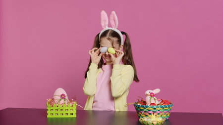 Enthusiastic young girl playing peek a boo in front of camera, using painted easter eggs against pink background. Joyful lovely toddler feeling excited about spring holiday festivity. Camera A.の写真素材