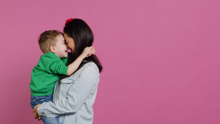 Adorable small boy hugging his mother and playing around, having fun against pink background. Young toddler posing with his mom holding him, laughing and being cheery. Camera A.の写真素材