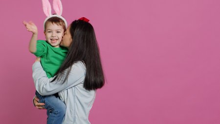 Mother lifting up her little boy and kissing him in studio, showing love and hugging cute kid against pink background. Sweet mom and son embracing each other and laughing. Camera A.の写真素材
