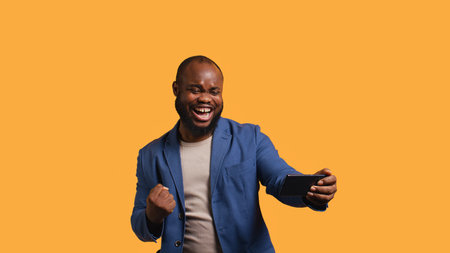 Happy man playing videogames on smartphone, celebrating after defeating foes, clenching hand gesture. Passionate BIPOC player thrilled about winning game on phone, studio background, camera Bの写真素材