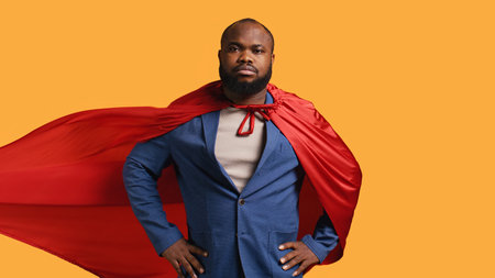 African american superhero flying with red cape, isolated over studio background, flexing muscles. Man wearing cloak posing as hero in costume showing courage and strength, camera Bの写真素材