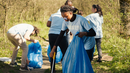 African american girl picking up trash with a long claw and garbage bags, cleaning forest habitat and fighting illegal dumping with a team of volunteers. Activist collecting rubbish. Camera A.の写真素材
