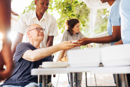 Charitable organisation gives assistance and free meals to homeless individuals, demonstrating dedication to fighting hunger and poverty. Volunteers provide a warm food to caucasian wheelchair user.の写真素材