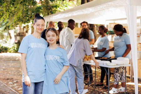 Portrait shot of a mother and daughter, supporting hunger relief initiative at an outdoor food bank. Two caucasian females wearing blue t-shirts written volunteer, ready to help the homeless people.の写真素材