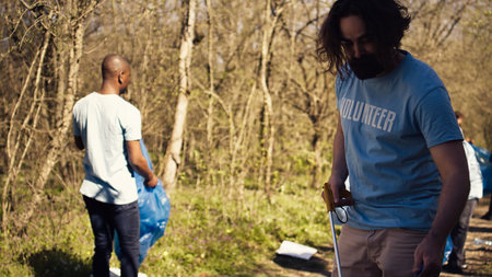 Man activist using tongs to grab garbage and plastic waste, picking up trash and cleaning the forest area. Volunteer sorting rubbish and recycling it, preserving the nature. Camera B.の写真素材