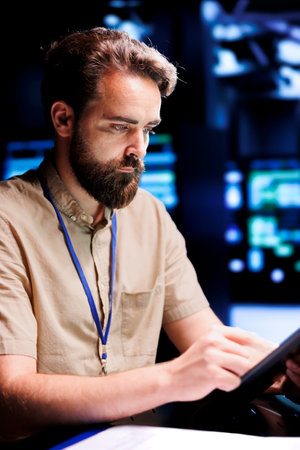 Repairman using tablet to examine server racks for power fluctuations, ensuring uninterrupted computing power. Worker monitoring data center, identifying potential vulnerabilitiesの写真素材