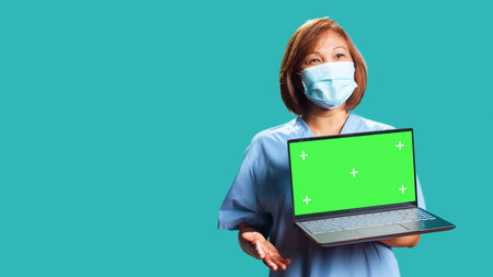 Close up handheld camera shot of happy clinic worker showing instructions video on laptop chroma key green screen. Nurse isolated over studio background presenting healthcare tape on gadgetの写真素材