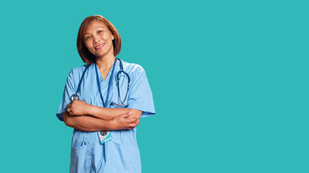 Joyful experienced asian nurse folding arms, smiling while looking at camera. Portrait of happy healthcare expert wearing protective scrubs, isolated over blue studio backgroundの写真素材
