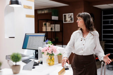 Elderly lady with suitcase lingers at front desk impatiently rings silver service bell to request help from staff with check-in. Caucasian old woman anxiously waiting to register at hotel reception.の写真素材