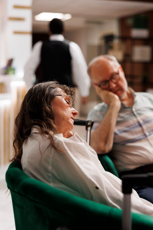 Detailed shot of retired senior woman and man, each sleeping on cozy couch while waiting to register for hotel room reservation. Photo focus on elderly female tourist resting in exclusive lounge area.の写真素材