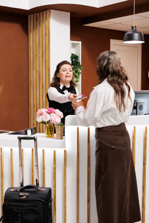Elderly female tourist checks in with her passport at hotel lobby desk. After confirming identity, friendly concierge at registration counter returns the personal documents of retired senior woman.の写真素材