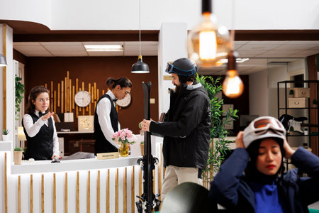 Female guest with helmet waits in luxurious lounge area ready for winter vacation at resort. Excitement fills the air as male traveler holding ski equipment checks in at hotel reception front desk.の写真素材