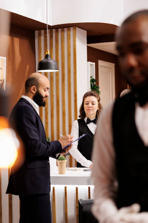 Businessman sign hotel records in lobby, doing check in upon his arrival at front desk reception. Receptionist providing luxury registration services, ensuring relaxing business trip for client.の写真素材