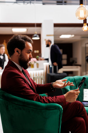 Businessperson on teleconference at hotel, sitting in lounge area with tablet discussing new partnership terms with CEO. Employee travelling for work purposes, videocall online.の写真素材