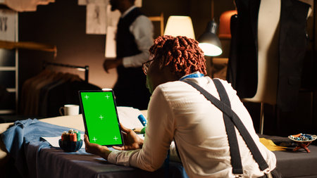Female tailor working with greenscreen on tablet, using isolated chroma key display in textile workshop. Woman holding device with blank copyspace on screen, handmade tailoring.の写真素材