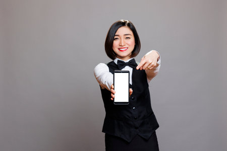 Smiling asian waitress holding smartphone and pointing with finger at blank white screen mockup and looking at camera. Receptionist showing mobile phone blank touchscreen portraitの写真素材