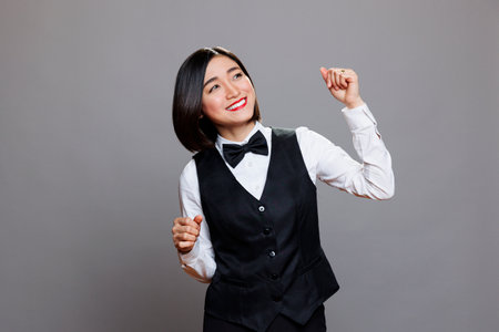 Attractive carefree asian woman receptionist wearing restaurant uniform joyfully dancing and having fun. Cheerful smiling young waitress moving arms to dynamic music beatsの写真素材