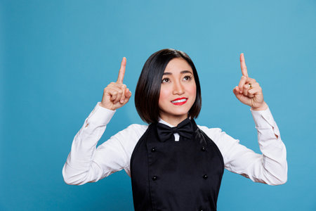 Cheerful smiling asian waitress in uniform pointing with fingers to up direction for catering service advertising. Restaurant woman worker showing upwards for product promotionの写真素材