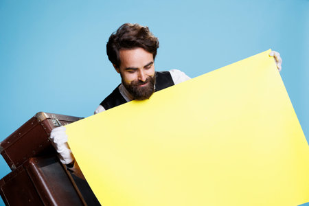 Hotel concierge holds empty yellow sign poses with blank banner on camera, showing isolated poster with empty copyspace for advertisement. Doorman with billboard ad sign over background.の写真素材